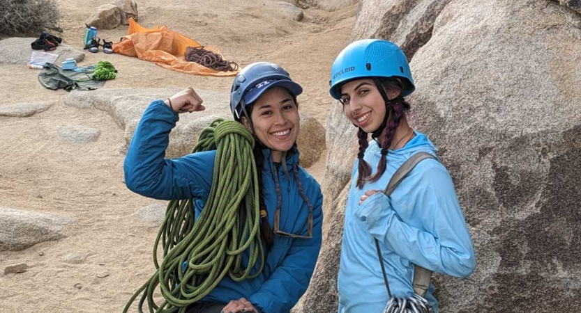 two people wearing helmets stand beside a large boulder and smile for the camera. They have climbing gear slung over their shoulders. 